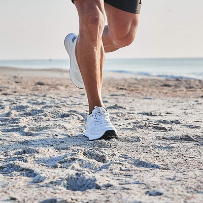 A person jogging on a sandy beach, wearing GORUCK's Men's Rough Runner shoes in light grey and dark grey for extra comfort, paired with black shorts. The ocean is visible in the background under a clear sky.