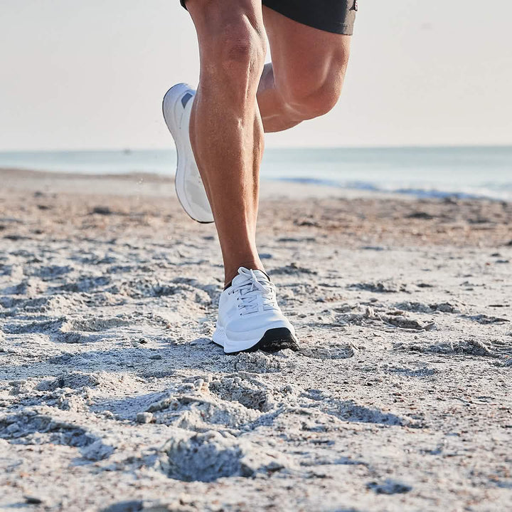 A person jogging on a sandy beach, wearing GORUCK's Men's Rough Runner shoes in light grey and dark grey for extra comfort, paired with black shorts. The ocean is visible in the background under a clear sky.