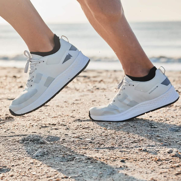 Close-up of a person running on a sandy beach, wearing GORUCK's Men's Rough Runner shoes in light grey and dark grey. The background features a blurred view of the ocean under a clear sky.