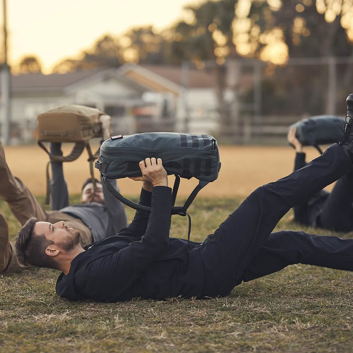 Three people exercise outdoors, lying on their backs and lifting weighted bags while wearing the Indestructible Challenge Windbreaker.