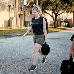 Woman walking outdoors carrying a black 25# GORUCK sandbag, wearing black GORUCK shirt, olive shorts, and backpack