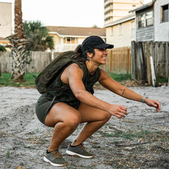 Fit woman wearing a black cap and camo tank top squatting outdoors with a GORUCK backpack in an urban setting