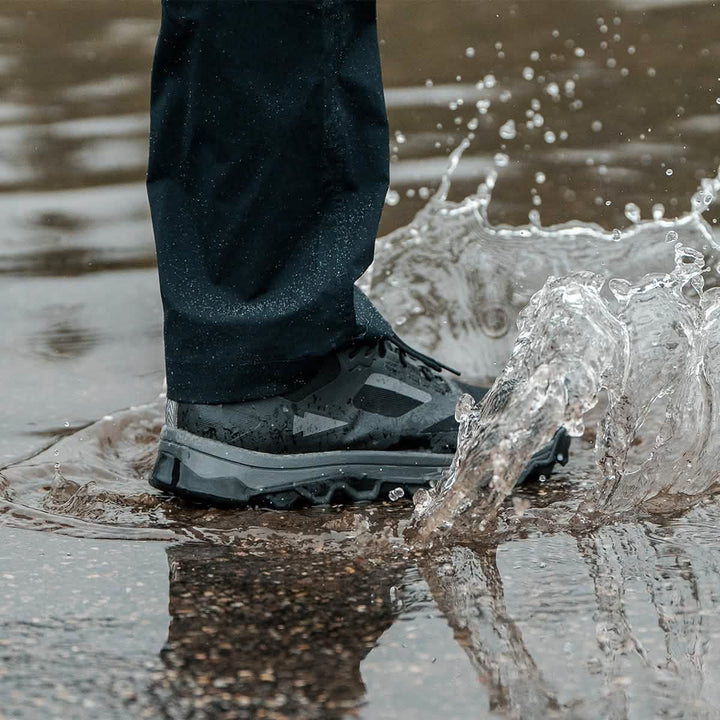 Someone wearing GORUCK Mackall shoes in Black + Forged Iron and dark pants is walking through a puddle. Water splashes around their feet, showcasing the shoes' water resistance. The wet and reflective surface makes it ideal for Special Forces training exercises.