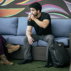 Man in black shirt and gray pants sitting on blue couch drinking from glass with GORUCK black backpack nearby