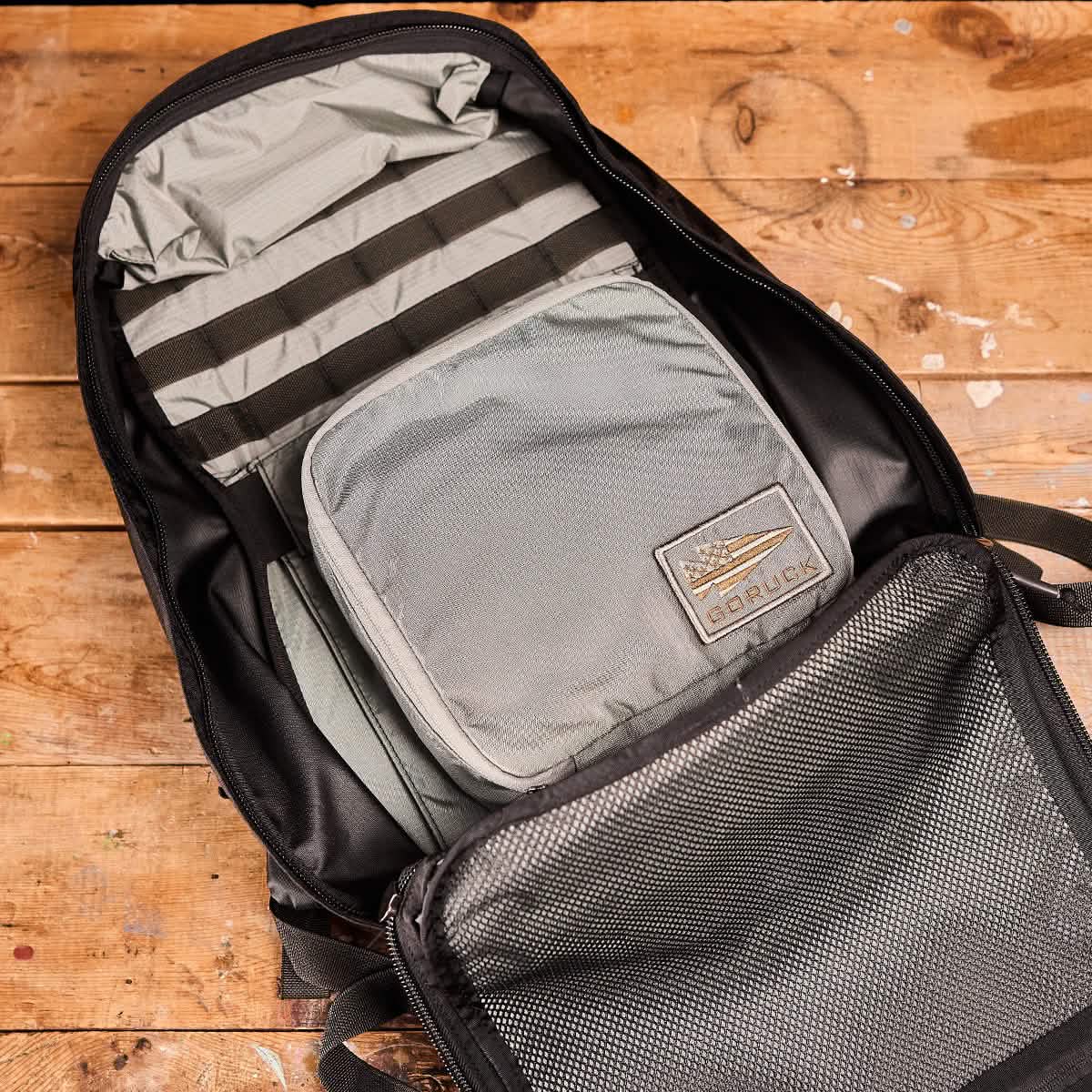 On a wooden surface, the backpack is displayed open, expertly organized with gray Shadow Pockets from GORUCK and featuring a branded patch. Constructed from durable 420D Ripstop ROBIC® Nylon, the bag's multiple compartments highlight efficient use of space within its neatly packed interior.