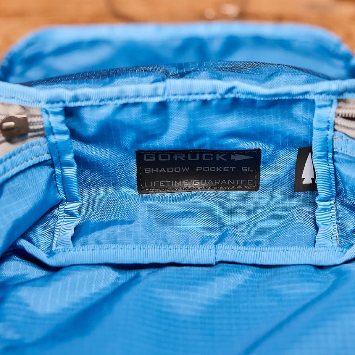 Close-up of a blue fabric interior of a backpack, showcasing a label that reads GORUCK Shadow Pockets - Ripstop ROBIC® and Lifetime Guarantee. The label, made with ROBIC Ripstop Nylon, is sewn near the zippered pocket's edge to optimize rucksack organization and ensure lightweight gear storage.