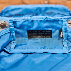 Close-up of a blue fabric interior of a backpack, showcasing a label that reads GORUCK Shadow Pockets - Ripstop ROBIC® and Lifetime Guarantee. The label, made with ROBIC Ripstop Nylon, is sewn near the zippered pocket's edge to optimize rucksack organization and ensure lightweight gear storage.