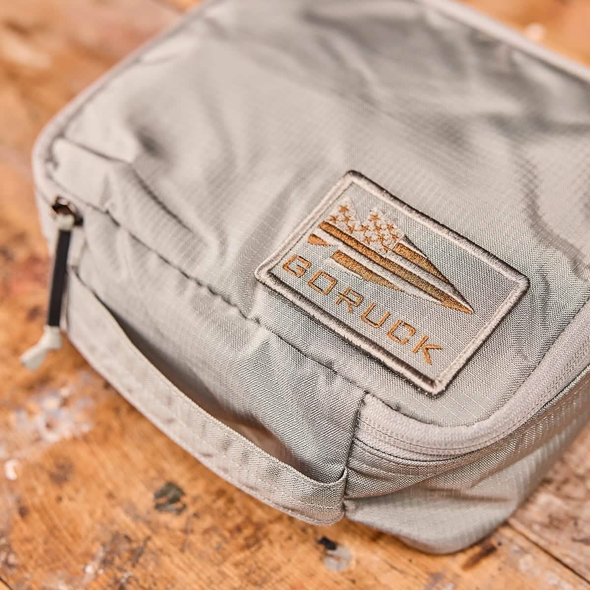 Close-up of a Shadow Pockets - Ripstop ROBIC® light gray fabric pouch by GORUCK, constructed from 420D ROBIC Nylon with a handle and zipper on a wooden surface. The pouch is adorned with an embroidered patch featuring a mountain and tree design, along with the word GORUCK, perfect for enhancing rucksack organization.