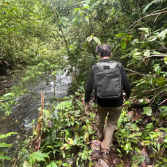 Hiker wearing GORUCK tactical black backpack walking along river in dense green jungle