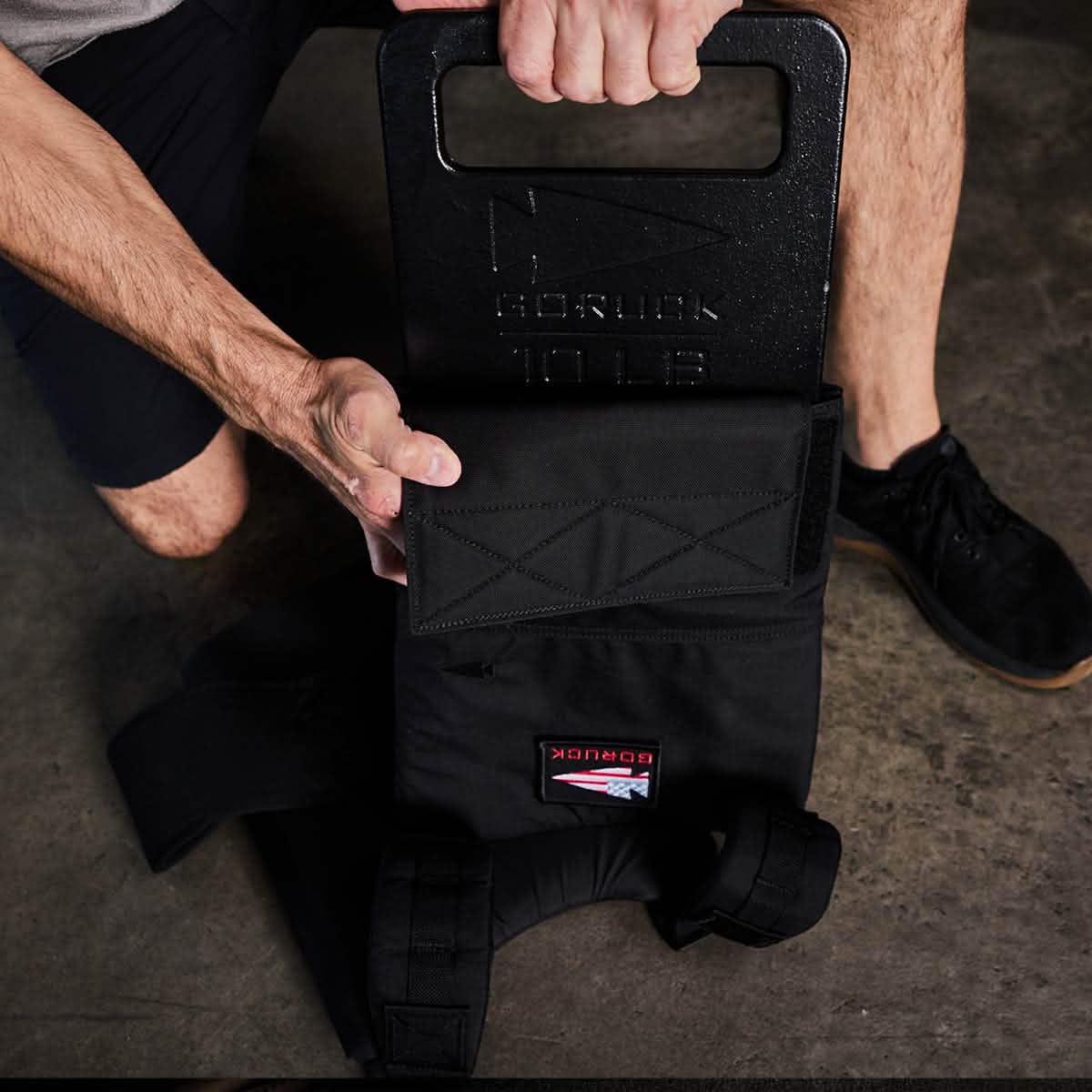 A person is kneeling on the concrete ground, holding a Rucker with a black rucksack prominently featuring a GORUCK Ruck Plate weighing 10 pounds. The rucksack has sturdy shoulder straps and displays a patch with red and white details, highlighting its American manufacturing quality.
