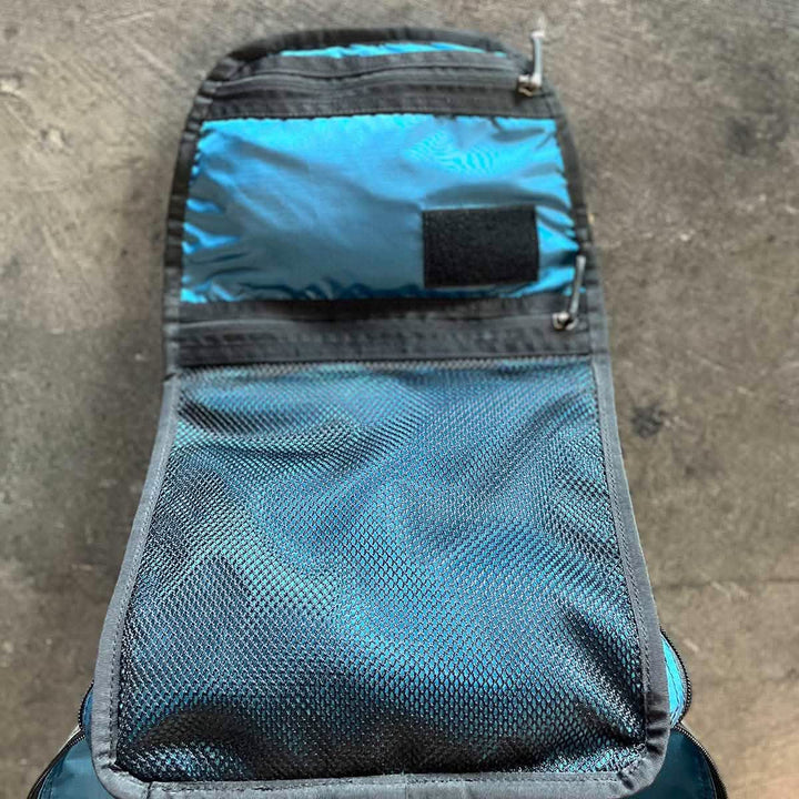The GORUCK Bullet 15L Robic Nylon - Built by SCARS toiletry bag, in blue and black, is made from durable Robic Nylon. It is open to show a mesh compartment and a small pocket with a Velcro closure. This bag sits on a textured, gray surface ready to complement your daypack essentials.