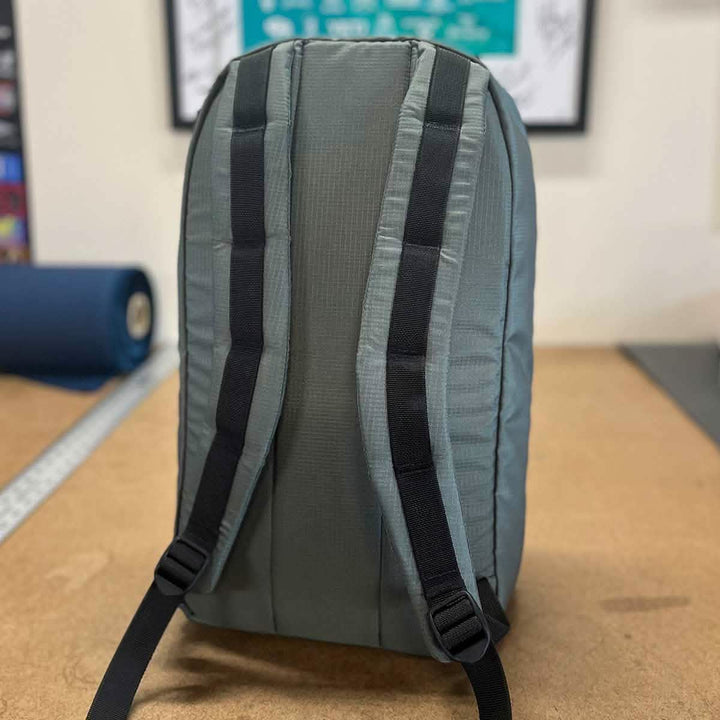 A Bullet 15L backpack from GORUCK's Built by SCARS collection, featuring gray Robic Nylon and black straps, stands upright on a wooden table. Designed for any adventure, it exudes durability and readiness. In the background, a blurred wall adorned with posters enhances its charm.