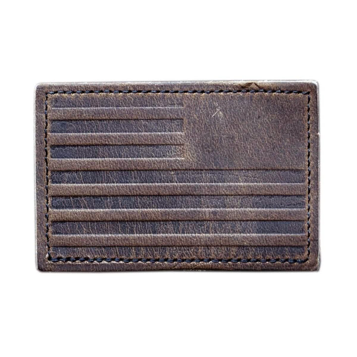 The Patch - Leather Reverse Flag by vendor-unknown is a rectangular piece crafted from genuine brown leather, featuring embossed horizontal lines and a square in the top left corner to create an abstract flag design. It has visible stitching along its edges and includes a discreet VELCRO® backing for easy attachment.