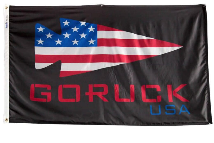 The Flag - GORUCK USA by Annin Flagmakers is expertly made from durable nylon. It features a black background with a spearhead design incorporating the American flag pattern. Beneath this, "GORUCK" is prominently displayed in bold red letters, with "USA" subtly written in smaller blue font.