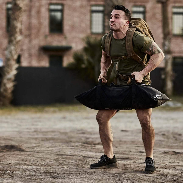 A man wearing a khaki backpack and a green t-shirt lifts a GORUCK Simple Training Sandbag, which features padded handles, outdoors. He stands on a dusty surface with a brick building and palm trees in the background, exerting effort as he performs the exercise.