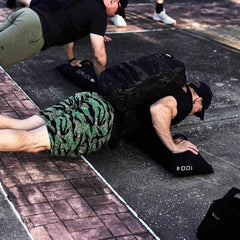 A person outdoors performs weighted push-ups with a camouflage backpack, likely filled with GORUCK's Simple Training Sandbags or gear from their home gym. Nearby, another individual mirrors the exercise. The ground is paved with bricks and asphalt. Both display dedication that reflects GORUCK's commitment to quality and persistence reminiscent of Scars Lifetime Guarantee.