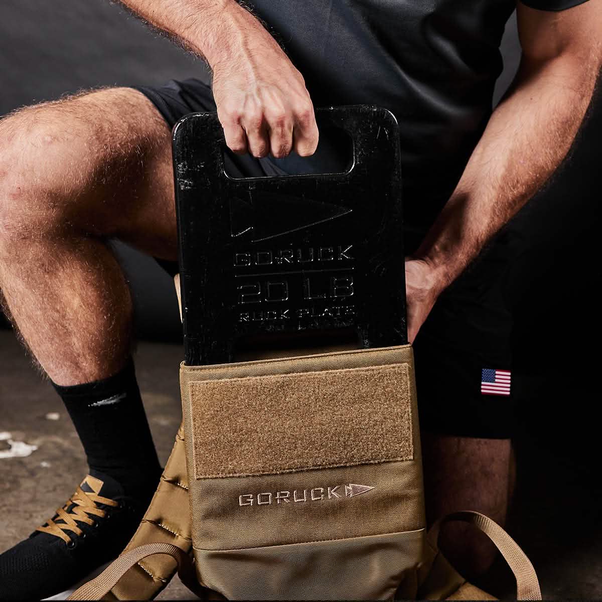 An individual is inserting a black 20 lb GORUCK Ruck Plate into a tan Rucker backpack. Dressed in a dark shirt and shorts adorned with a small American flag patch, the person highlights the excellence of American craftsmanship. This scene takes place indoors on a concrete floor.