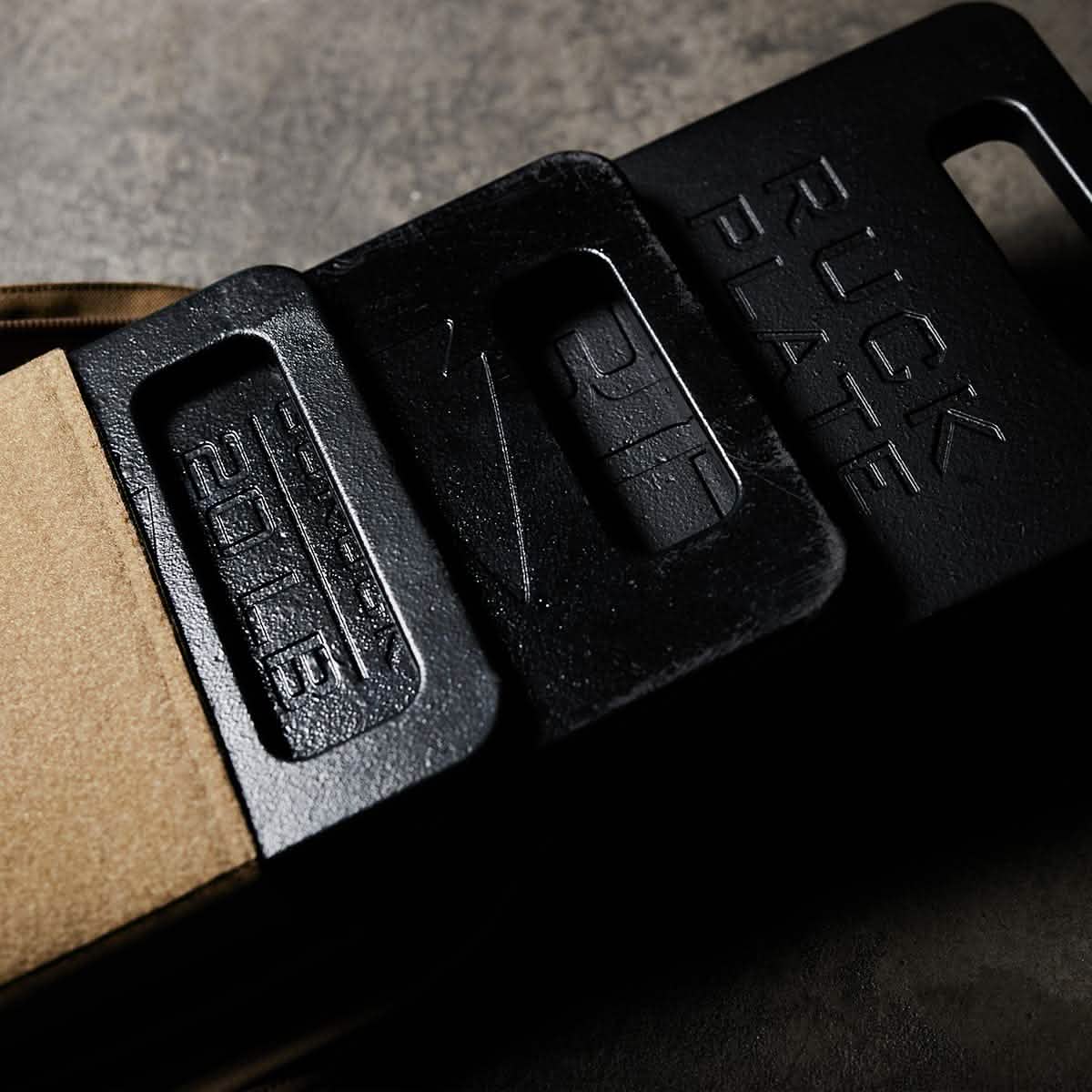 Close-up of two overlapping GORUCK Ruck Plates made from black metal with embossed text. The top plate features 'RUCK PLATE', and the bottom one is marked '20LB'. These American-made products lie on a textured gray surface, partially covering a cardboard box.