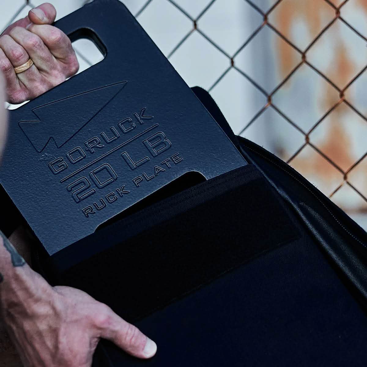 A person inserts a GORUCK Ruck Plate, weighing 20 lbs and featuring a rectangular cutout handle, into a Rucker. This American-made black ruck plate is prominently displayed against the wire fence background.
