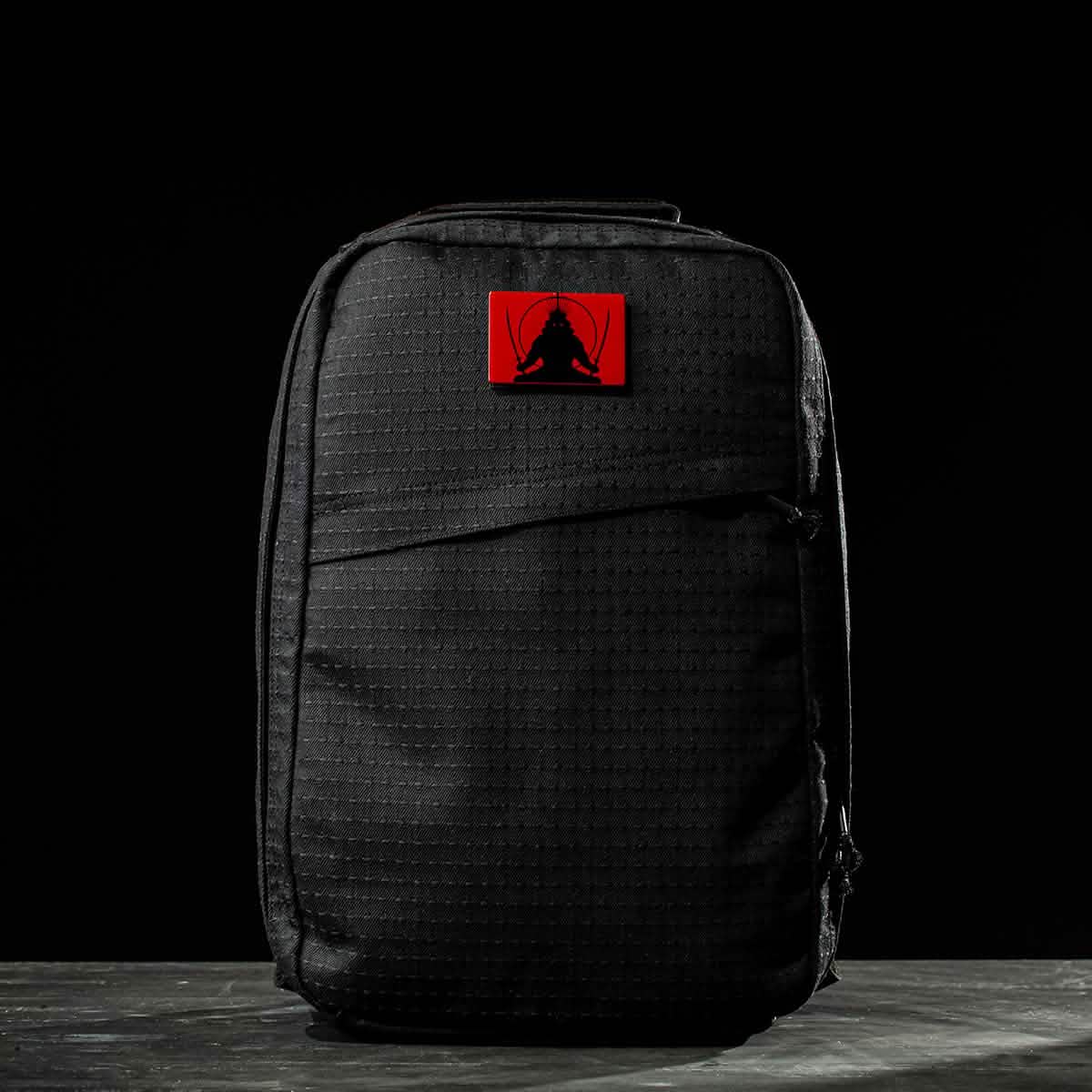 A GR1 USA x Carryology - GRXC2 Samurai backpack in black, featuring a red patch with a seated figure and made from premium Japanese cotton, set against a dark background.