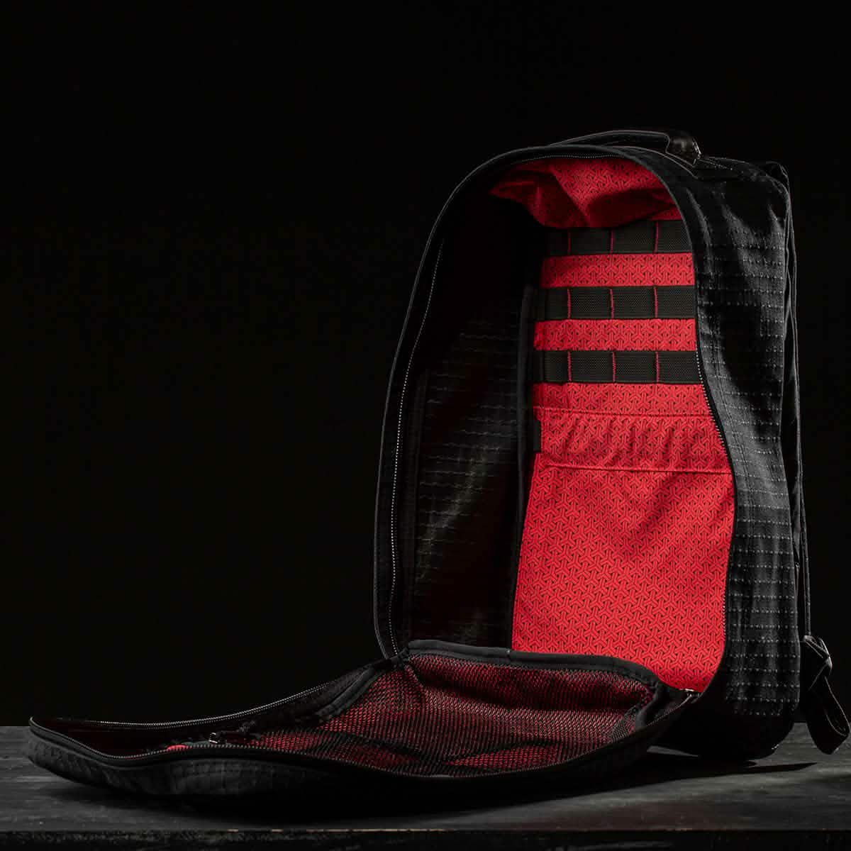 The GR1 USA x Carryology - GRXC2 Samurai is a black backpack with a red interior, unzipped and upright against a dark background, crafted from Japanese cotton—highlighting the signature detail of this exclusive collaboration.