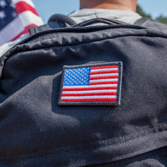 A person with a backpack adorned by the vendor-unknown "Patch - American Flag," showcasing the flag prominently in the center. The pride for the US of A is evident as an American flag is also partially visible in the background.