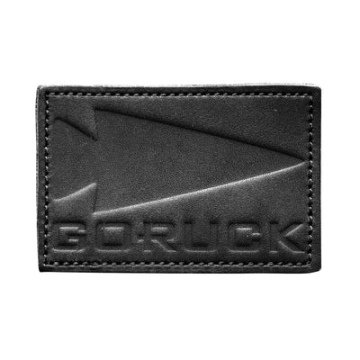 The Patch - Leather GORUCK Spearhead from vendor-unknown is made from black full-grain cowhide leather and features an arrow embossed alongside the text "GORUCK." It has visible stitching along the edges, a slightly textured surface, and comes with a VELCRO® backing for easy attachment.