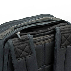 A close-up of a black GORUCK GR1 USA - Heritage rucksack highlights the padded shoulder straps and a slightly open top zipper offering a glimpse into its interior. Made from waxed duck canvas, the stitching on both the material and straps is prominently visible.