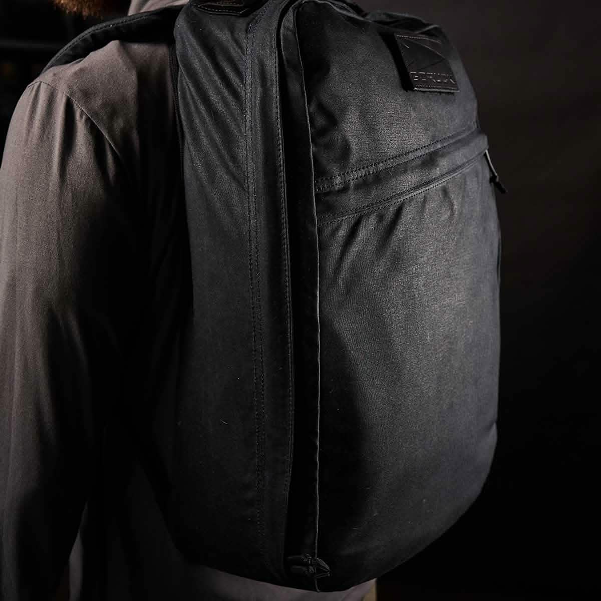 A person wearing a dark shirt is shown from the back, carrying the GORUCK GR1 USA - Heritage backpack, crafted from waxed duck canvas. The dimly lit setting accentuates the texture and shape of this heritage rucksack, highlighting its durability and timeless appeal.