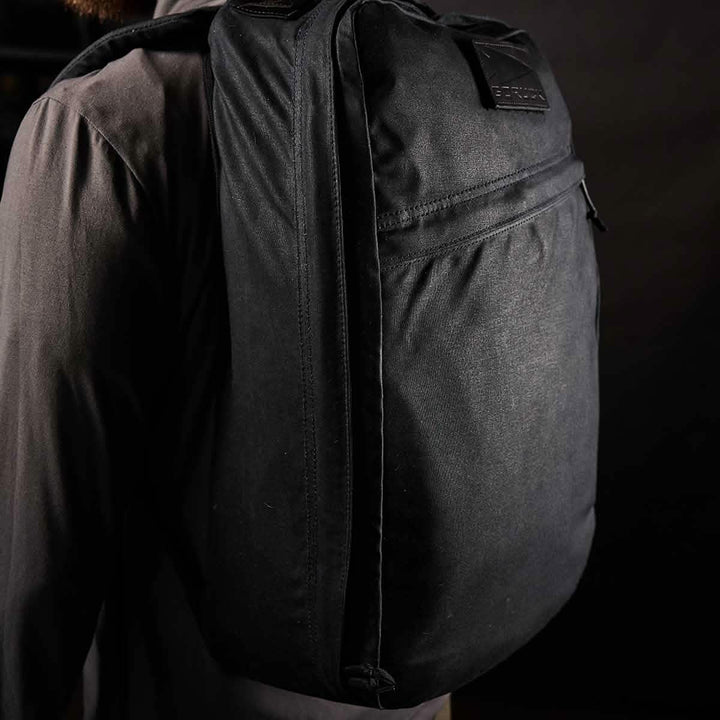 A person wearing a dark shirt is shown from the back, carrying the GORUCK GR1 USA - Heritage backpack, crafted from waxed duck canvas. The dimly lit setting accentuates the texture and shape of this heritage rucksack, highlighting its durability and timeless appeal.