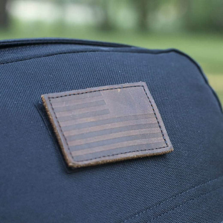 Close-up of a black fabric bag showcasing a rectangular patch with a matte, monochromatic reverse American flag design. The patch has a VELCRO® backing and is from the brand vendor-unknown. The background features blurred green foliage.