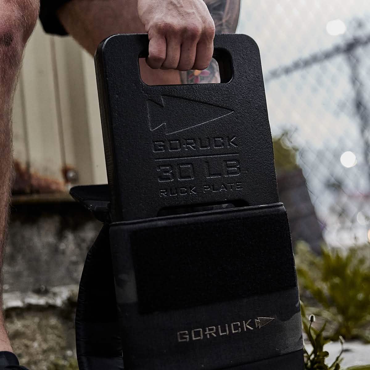 Next to a chain-link fence, someone places a 30 lb black GORUCK Ruck Plate into their Rucker backpack. The scene highlights the quality of American manufacturing, with the GORUCK logo prominently featured on both the plate and the backpack.