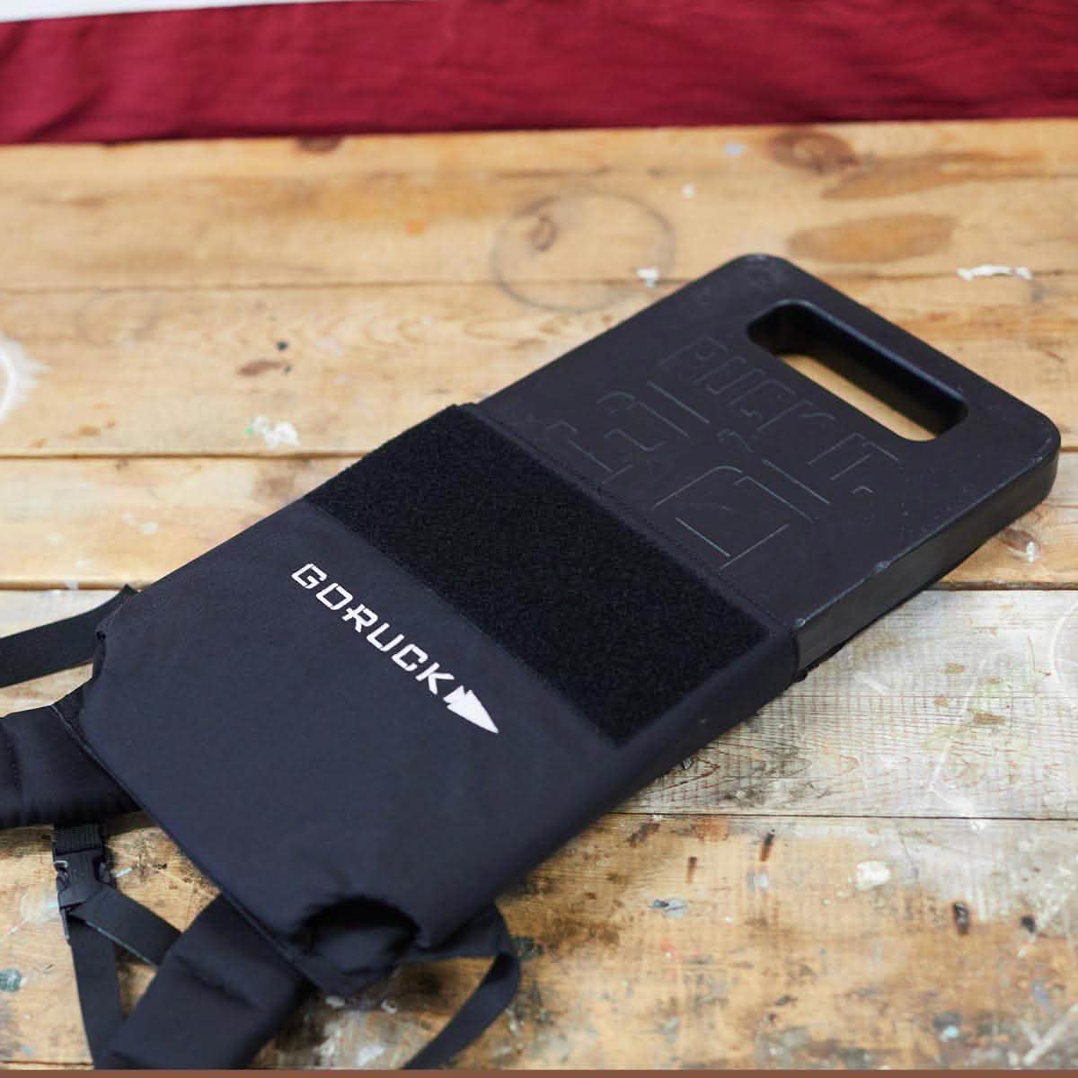 A black GORUCK Rucker backpack sits on a wooden surface, partially obscuring an American flag in the background. Highlighting the superior quality of American craftsmanship, this bag includes a velcro patch area and a durable handle, designed to effortlessly accommodate your Ruck Plates.