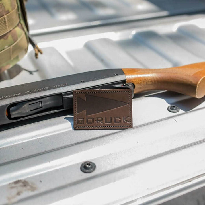 A shotgun with a wooden stock rests on a metal surface, adorned with the "Patch - Leather GORUCK Spearhead" by vendor-unknown. Crafted from full-grain cowhide, this patch exudes rugged charm. In the background, a camouflaged backpack is partially visible.