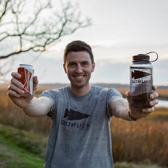 An outdoorsman with a smile holds a Budweiser can in one hand and a 32oz Nalgene Water Bottle featuring the GORUCK Spearhead design in the other. He's dressed in a gray GORUCK t-shirt, set against a backdrop of grassy fields, trees, and a partly cloudy sky.
