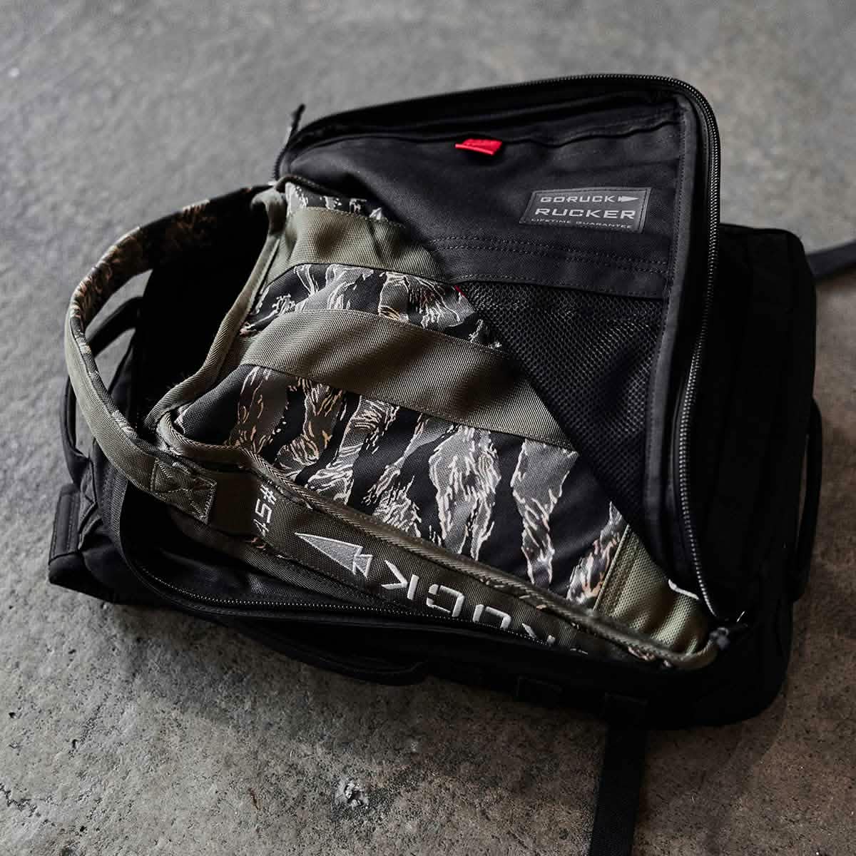 A Sand Jerry Can by GORUCK, crafted from Special Forces Grade materials, stands partially unzipped to reveal its camouflage-patterned interior and a gray and white Rucker patch. It rests on a textured gray surface, showcasing both toughness and style.
