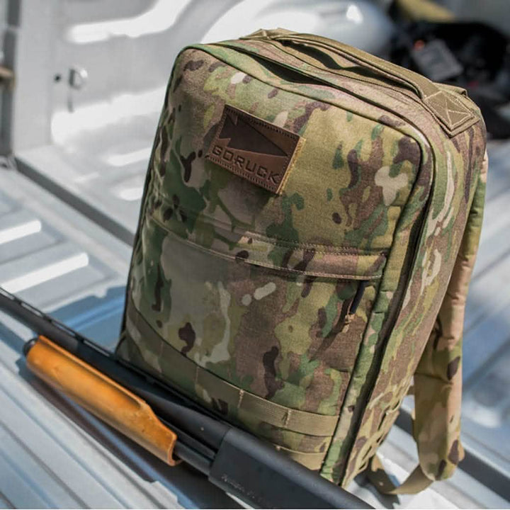 A camouflage backpack adorned with a logo on the strap rests on a metallic surface, its design highlighted by the Leather GORUCK Spearhead patch. In front of it, against a gray, textured backdrop, lies a wooden-handled tool or weapon.