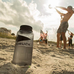 A robust Nalgene Water Bottle (32oz) - GORUCK Spearhead is placed in the sand on a sunlit beach. In the distance, a group engages in exercise, with some members lifting kettlebells while an American flag waves gently beneath a partly cloudy sky.