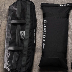 Two Sandbags 2.0 by GORUCK, crafted from durable black 1000D CORDURA and featuring white logos, are placed on a textured surface. The larger training sandbag with multiple handles is positioned on the left, while the smaller one lies flat on the right. Both are backed by the trusted Scars Lifetime Guarantee.