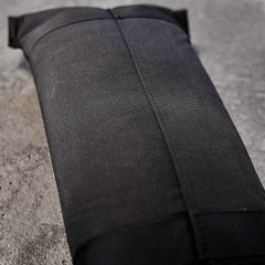A black rectangular GORUCK Sandbags 2.0 rests on a textured gray surface. Made from durable 1000D CORDURA fabric, it showcases a visible central seam and reinforced edges for added strength. Backed by the Scars Lifetime Guarantee, this robust sandbag is designed to endure even the most intense workouts.