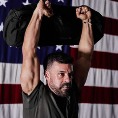 A person is hoisting a Sandbags 2.0 by GORUCK above their head, highlighting their muscular arms. They stand in front of an American flag, donned in a dark shirt, with a determined look on their face, epitomizing strength and resilience.