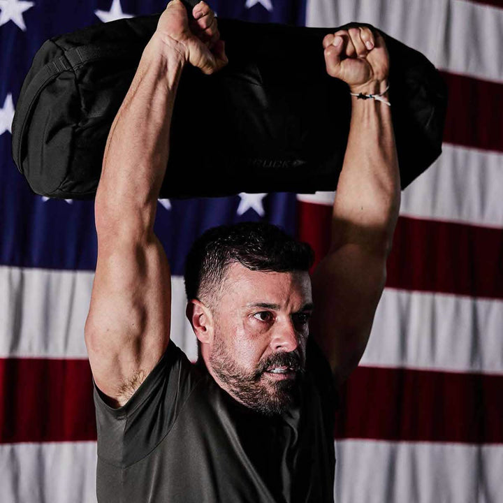A person is hoisting a Sandbags 2.0 by GORUCK above their head, highlighting their muscular arms. They stand in front of an American flag, donned in a dark shirt, with a determined look on their face, epitomizing strength and resilience.