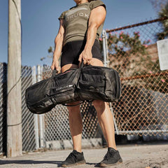 Against an urban backdrop with clear skies, a person in a green T-shirt and black shorts lifts the GORUCK Sandbags 2.0, featuring heavy-duty handles. The scene is framed by a fence and utility pole, highlighting strength and determination amidst the city's pulse.