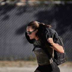 A person wearing sunglasses is running outdoors, carrying the Sandbags 2.0 by GORUCK with heavy duty handles over their shoulder. The background features a wire fence and some trees, with sunlight casting shadows.