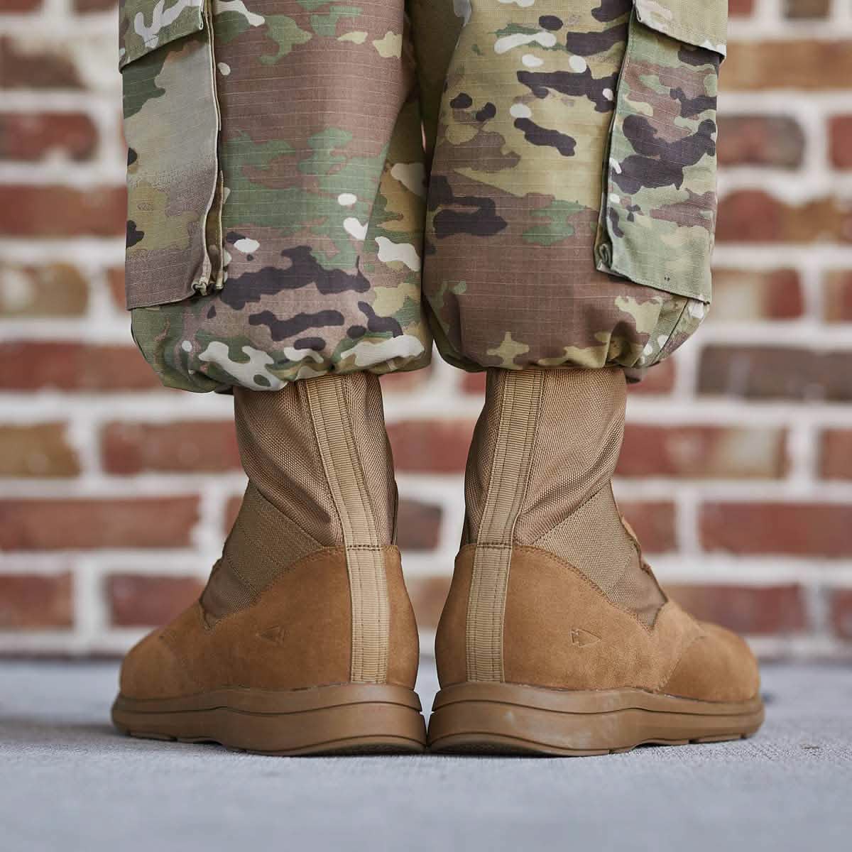 A close-up of a person wearing camouflage pants and MACV-1 High Top Coyote Suede boots by GORUCK, standing on a concrete surface. A brick wall is visible in the background.