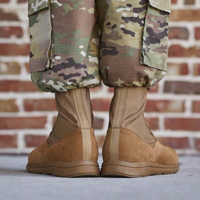 A close-up of a person wearing camouflage pants and MACV-1 High Top Coyote Suede boots by GORUCK, standing on a concrete surface. A brick wall is visible in the background.