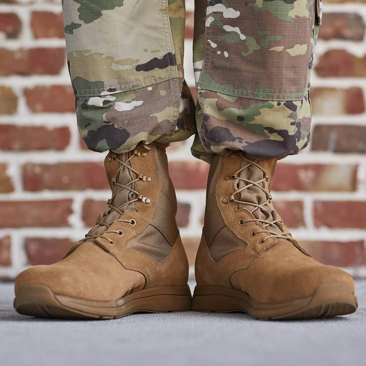 A close-up of a person wearing MACV-1 - High Top boots by GORUCK, compliant with AR 670-1 standards, paired with camouflage pants on a concrete surface. The coyote suede military boots showcase a durable all-terrain outsole and suede leather upper. A red brick wall serves as the background, completing the rugged look.