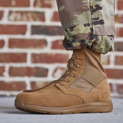 A close-up shows a person wearing camouflage pants tucked into GORUCK's MACV-1 High Top boots in Coyote Suede, featuring an all-terrain outsole, while standing on a concrete surface with a brick wall in the background.