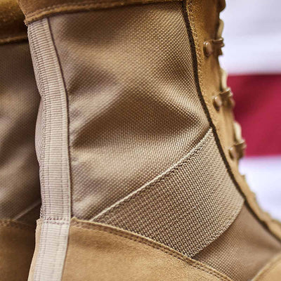 Close-up of the MACV-1 High Top by GORUCK, a tan military-style boot featuring textured fabric and reinforced stitching, with a Coyote Suede leather upper that enhances its rugged durability. The background displays hints of red and white stripes, highlighting the boot's all-terrain outsole.
