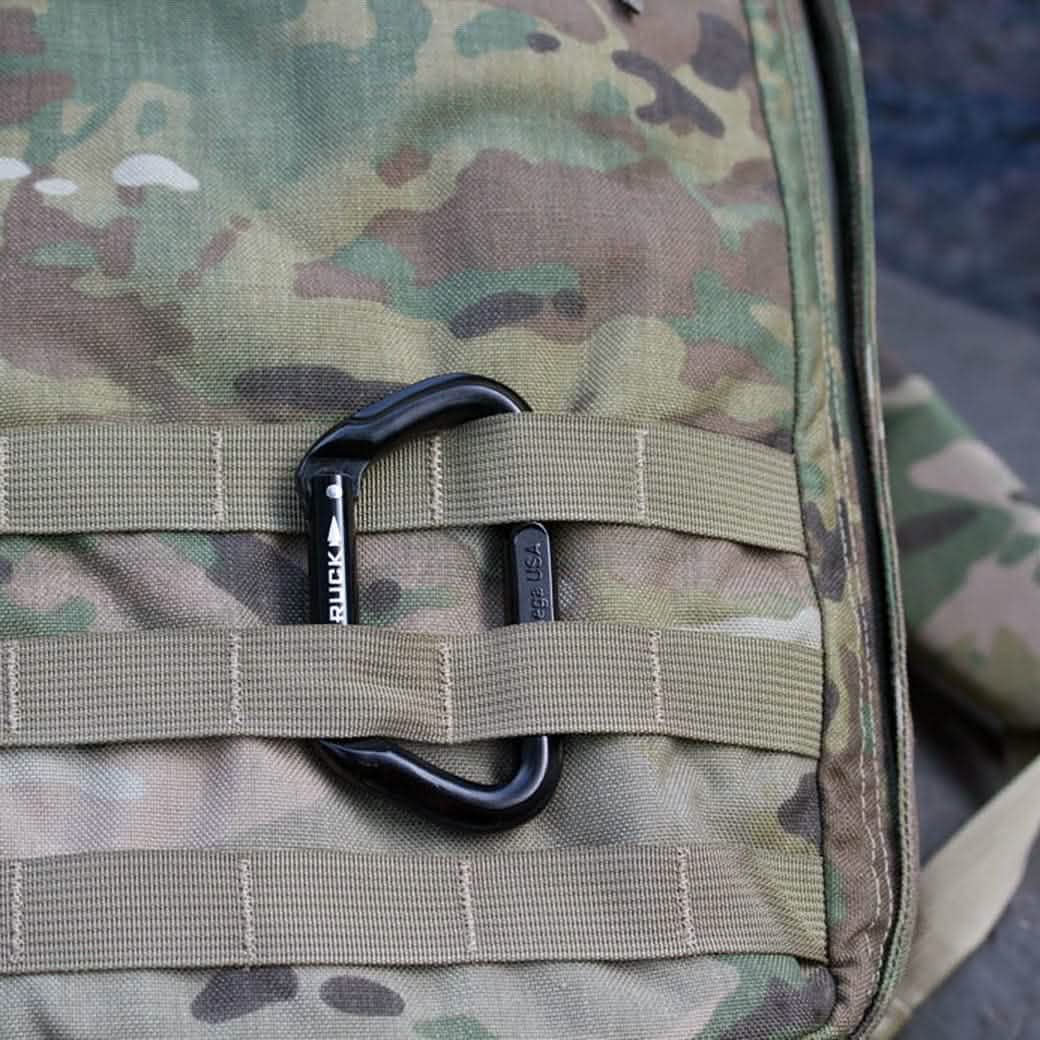 Close-up of a camouflage fabric showcasing the SMC Gear Carabiner with GORUCK Logo, attached to beige MOLLE webbing. The pattern, featuring shades of green, brown, and beige, is typically utilized in military and outdoor gear for effective concealment.
